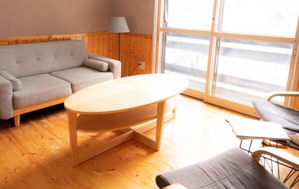 Una habitación con piso de madera, un sofá gris, una mesa de madera y una ventana con vistas.