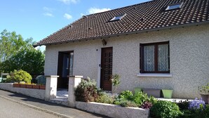 Ein zweistöckiges Haus mit Ziegeldach, einer Haustür und Fenstern. Vor dem Haus befinden sich Kübelpflanzen und ein kleiner Garten.