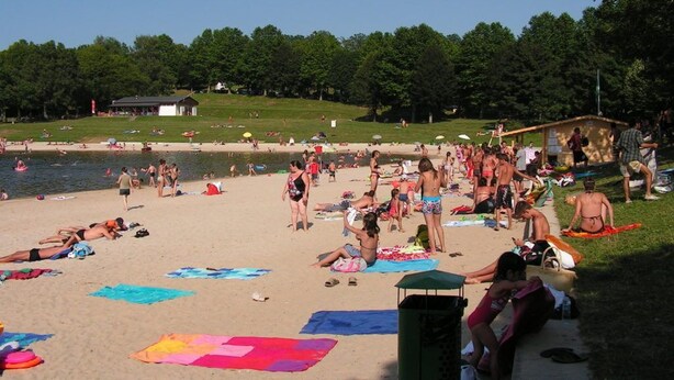Ein Strand mit badenden und sonnenden Personen, eine begrünte Fläche mit einem Gebäude und einem klaren Himmel.