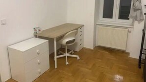 Une petite pièce, bien rangée, avec un plancher en bois, un bureau blanc doté de tiroirs, une chaise blanche et un radiateur.