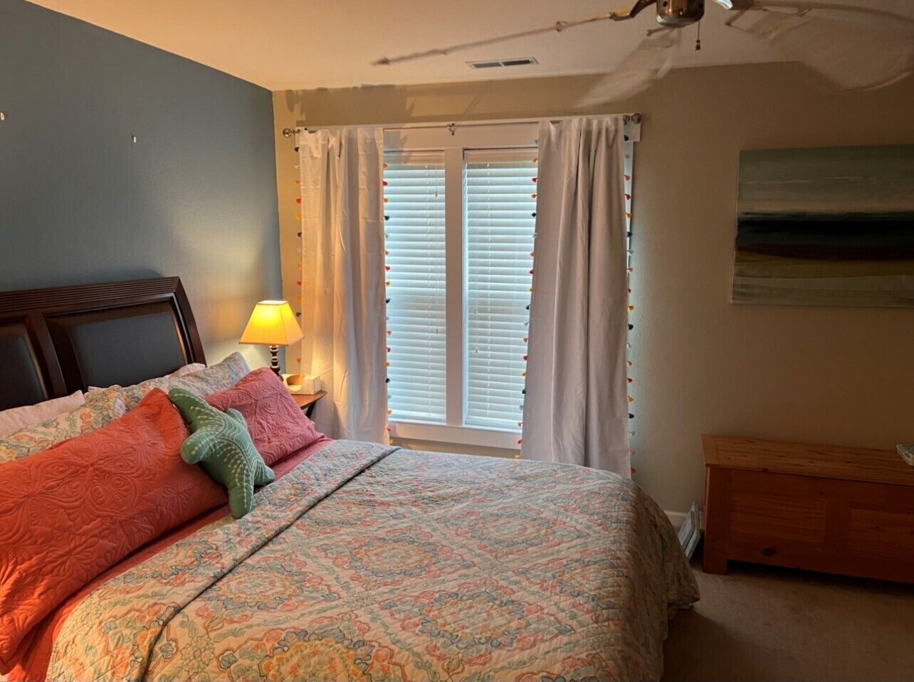 Une chambre à coucher comprenant un lit, une table de chevet avec une lampe, une fenêtre avec des stores et une commode en bois.