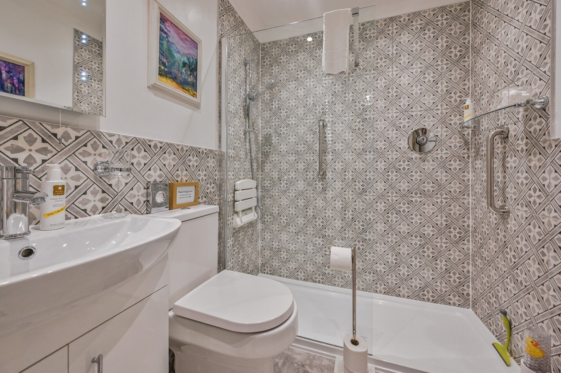 Ein modernes Badezimmer mit einem weißen Waschbecken, einer Toilette und einer Badewanne. Die Wände sind mit einem geometrischen Muster gefliest. Ein gerahmtes Bild hängt an der Wand und ein Handtuch an der Tür.