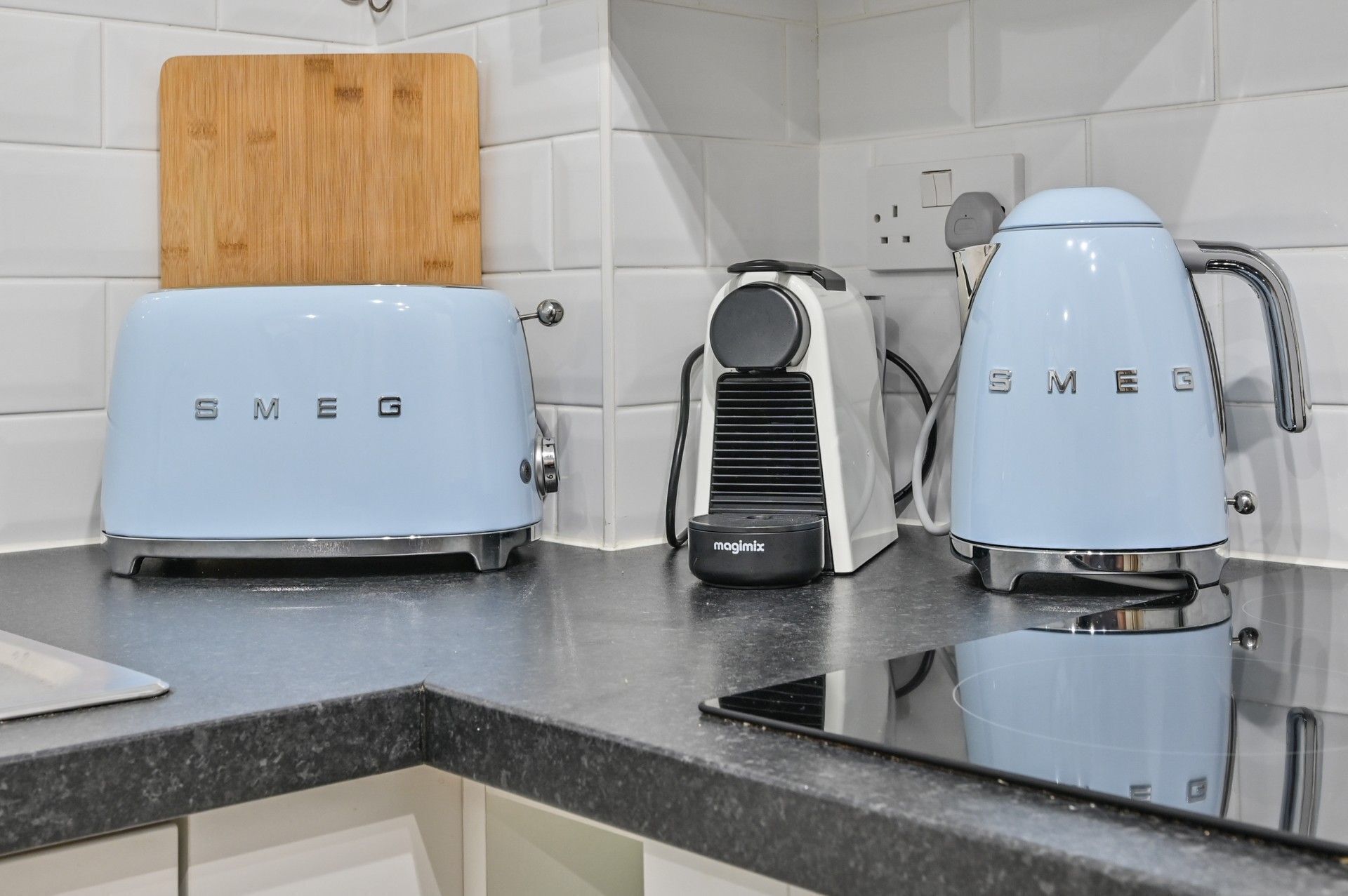 A kitchen with two light blue Smeg appliances, a wooden cutting board, and white tiled walls.