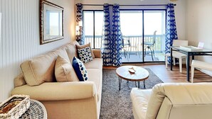 A living room with a beige sofa, a round coffee table, and a white armchair. There is a balcony with blue curtains and a ceiling fan. - Beach Racquet A221 by Tybee Vacation Rentals (Tybee Island)