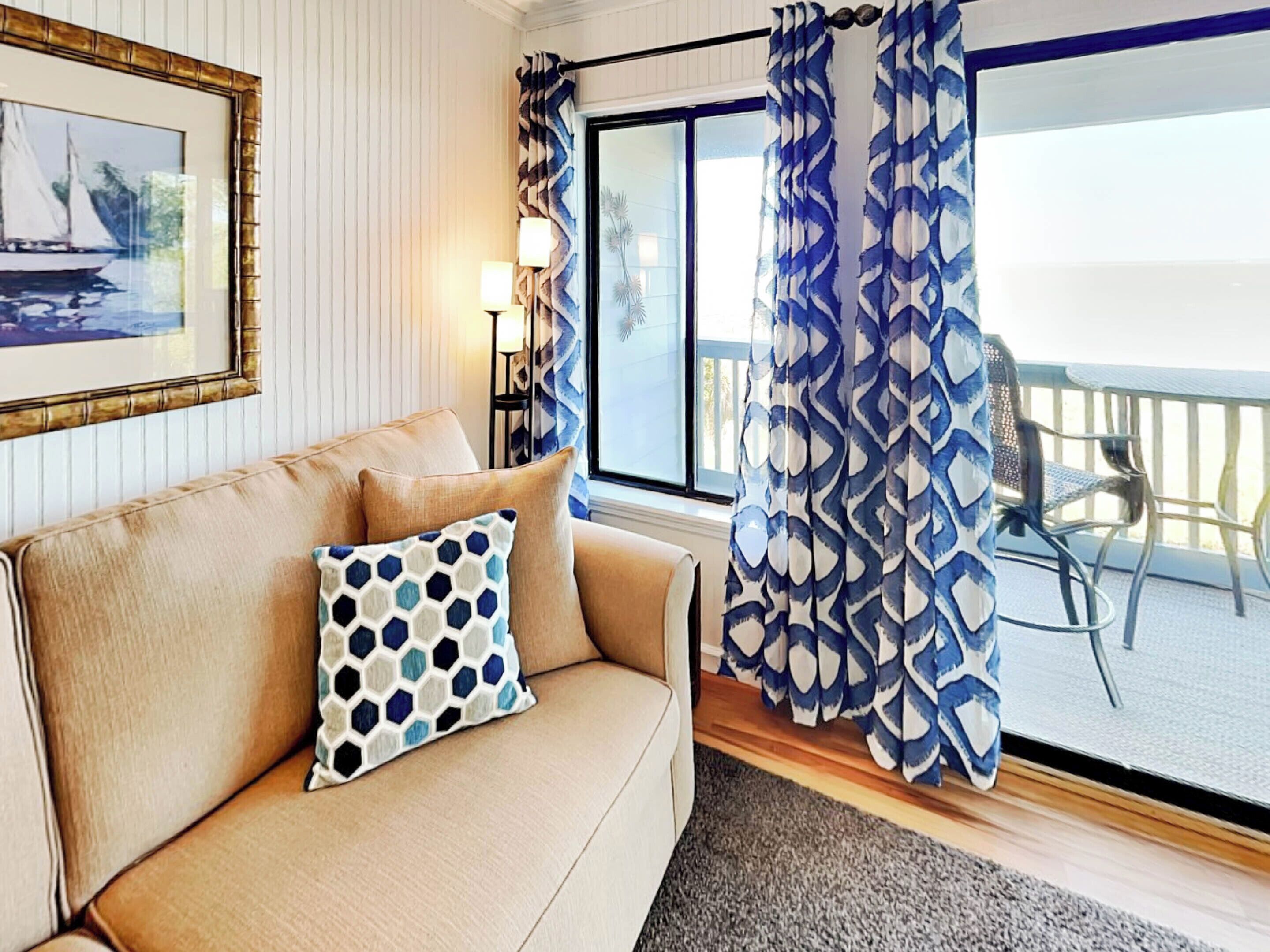 A living room with a beige sofa, a patterned pillow, a framed picture on the wall, and a view of the balcony with patterned curtains.