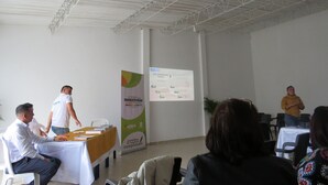 Un salón de conferencias con una pantalla de presentación, una mesa con documentos y asistentes sentados en mesas.