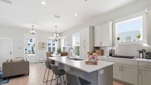 A modern kitchen with white cabinetry, a central island with bar stools, a large window, and a chandelier. - The Way Of Water 3 Bedroom Home by RedAwning (Panama City Beach)