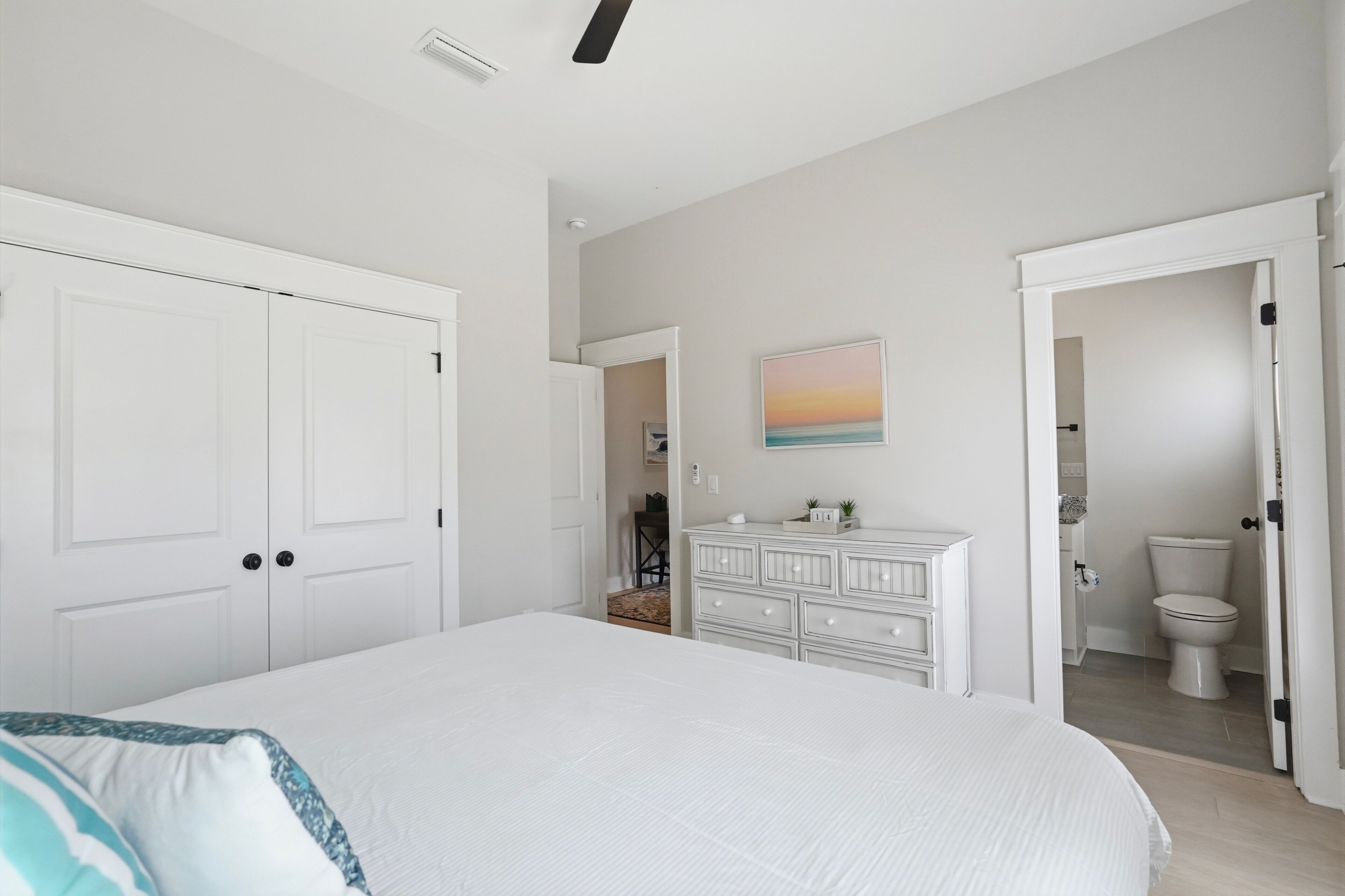 A bedroom with a large bed, a dresser, a painting on the wall, and a bathroom visible through an open door.