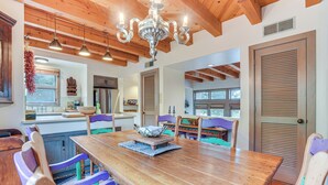 Uma sala de jantar ampla com mesa de madeira e cadeiras coloridas, um lustre e vigas de madeira no teto.
