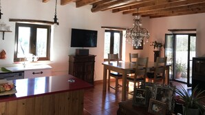 A spacious kitchen with wooden beams, a dining area with chairs and a table, a TV mounted on the wall, and a chandelier. - Casa da Oliveira (Pedrógão Grande)