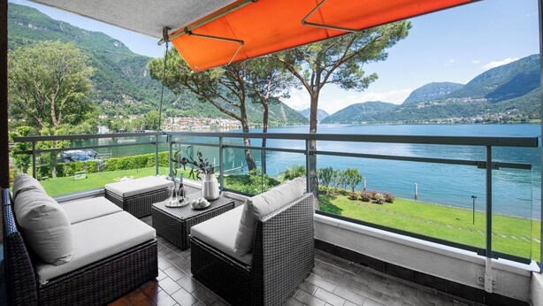 Un balcon agrémenté de meubles en osier et d’un parasol orange, offrant une vue sur un lac et des montagnes.