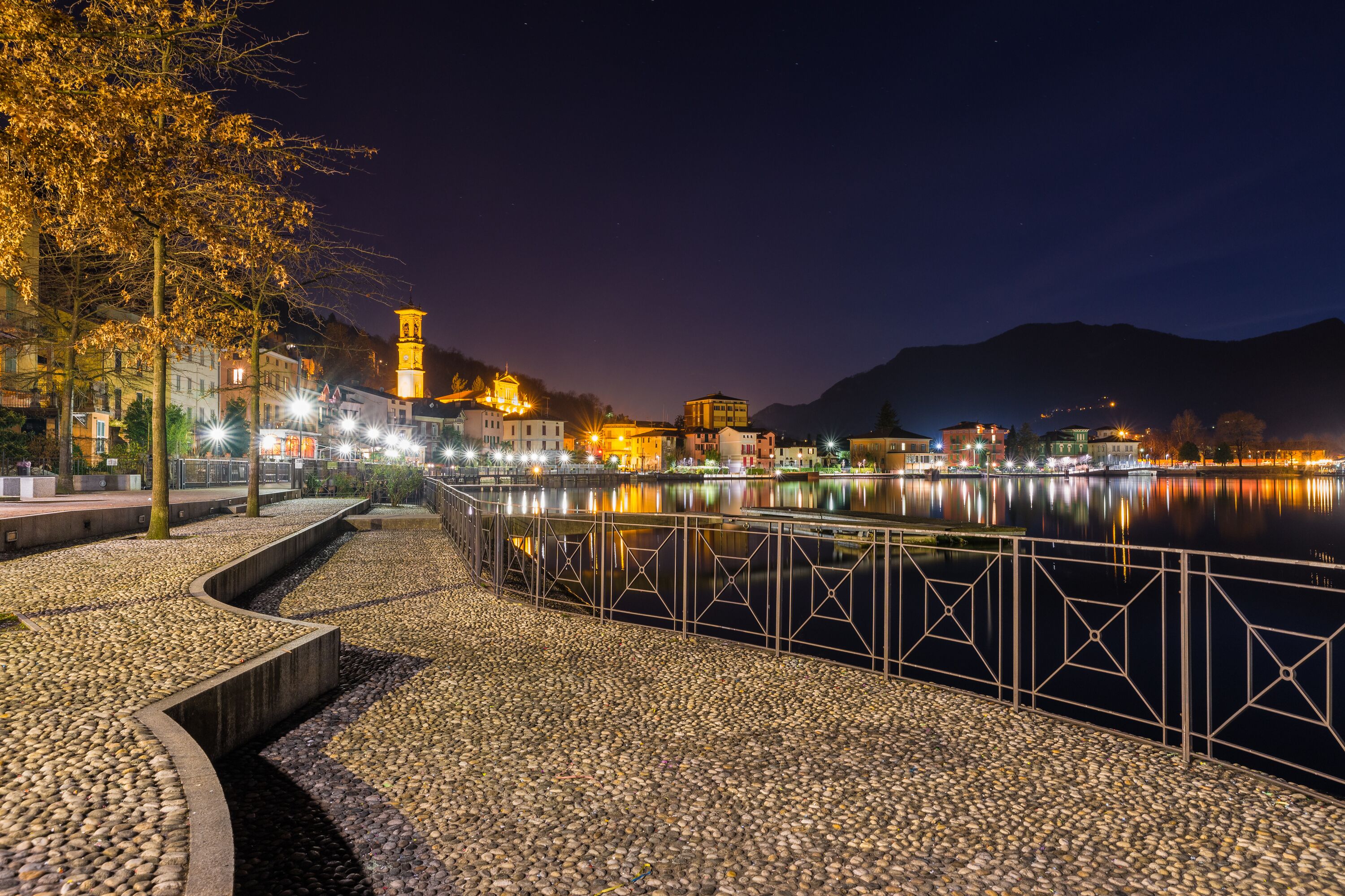 A nighttime view of a riverside promenade with a stone pathway, metal railing, and illuminated buildings reflecting on the water.