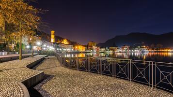 A nighttime view of a riverside promenade with a stone pathway, metal railing, and illuminated buildings reflecting on the water.
