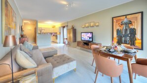 Un salon moderne avec un canapé, un pouf, une table à manger et des chaises. Un téléviseur est fixé au mur, et un tableau est accroché au-dessus de la table.