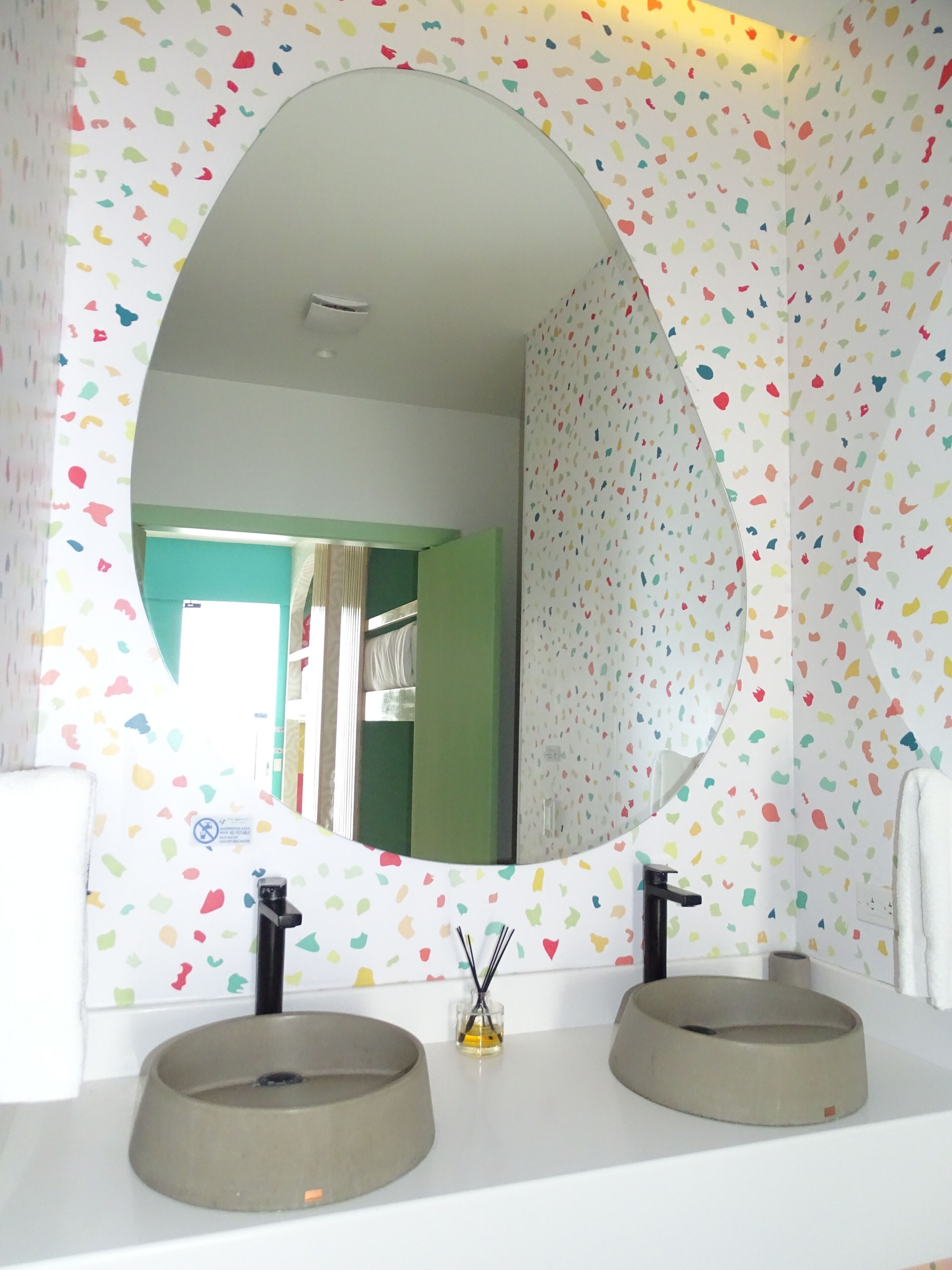 Un baño con un espejo redondo, dos lavamanos y una pared con un estampado de hojas de colores.