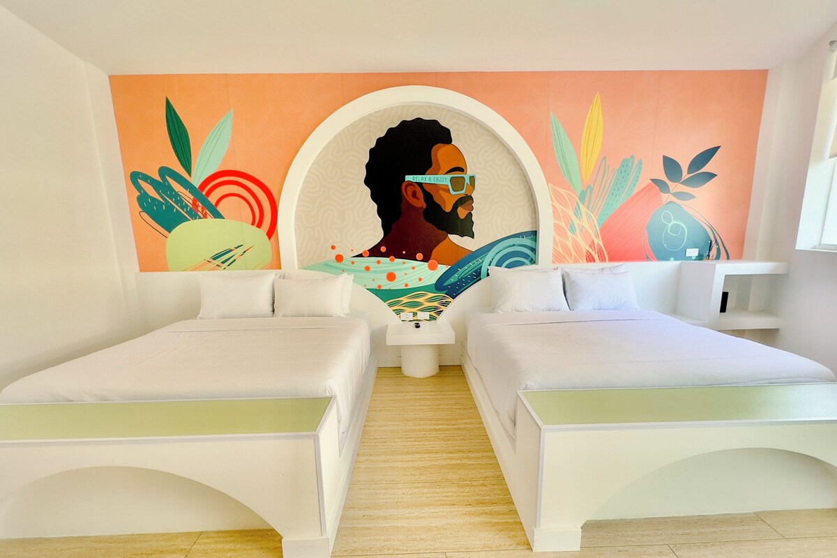 A modern hotel room with two white beds, a mural on the wall, and a small white table.