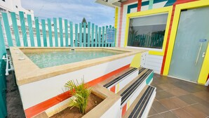 Una piscina con solárium de azulejos, un pequeño jardín y un edificio colorido que indica una zona de natación.