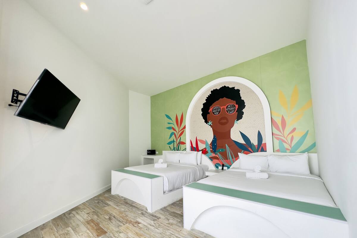 Habitación de hotel con dos camas, un televisor de pantalla plana montado en la pared y un mural colorido.