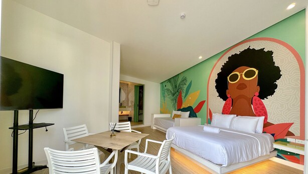 Una habitación de hotel moderna con una cama, una mesa de comedor con sillas, un televisor de pantalla plana y un mural colorido en la pared.