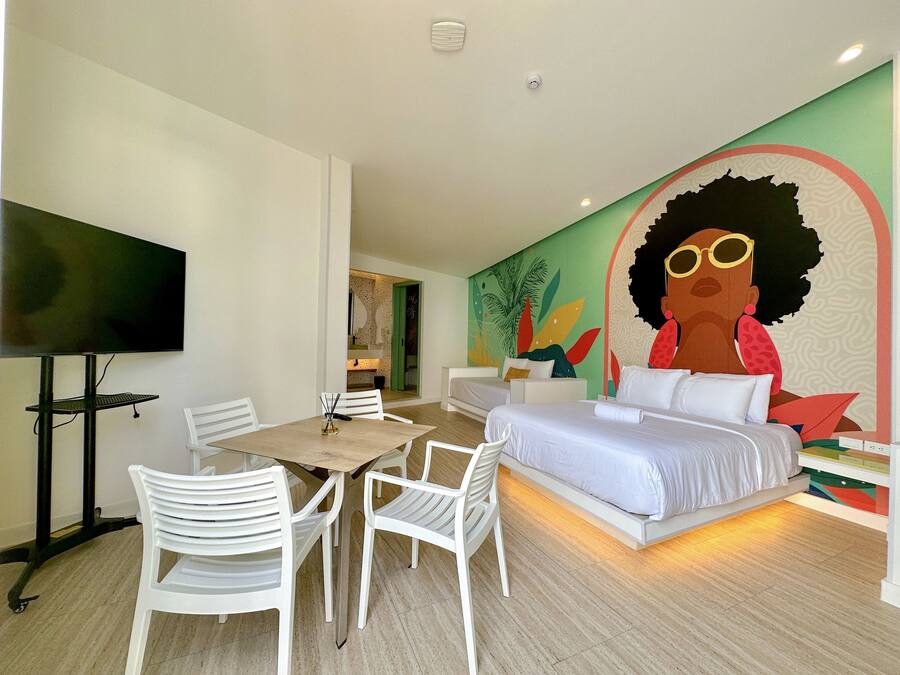 A modern hotel room with a bed, a dining table with chairs, a flat-screen TV, and a colorful mural on the wall.