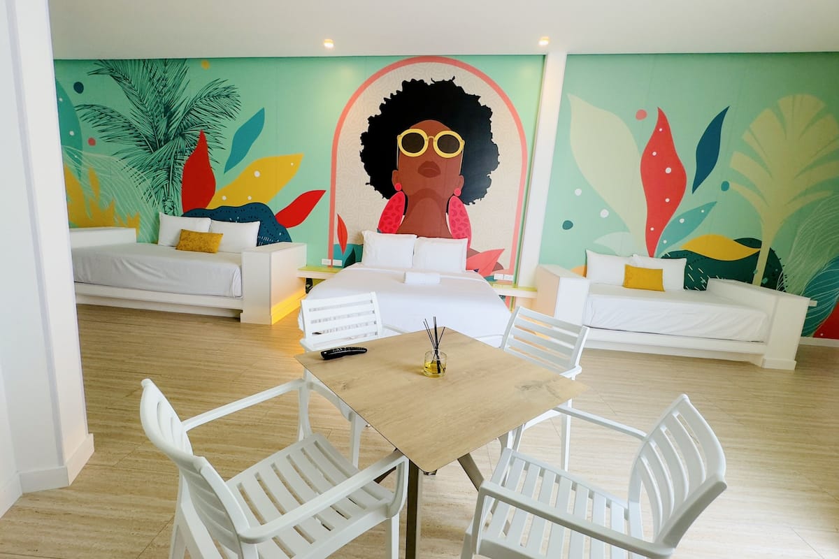 Una habitación de hotel moderna con un gran mural, dos sofás blancos, una mesa de madera y sillas blancas.