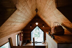 A cozy attic room with wooden beams, a hanging light fixture, a window with a view of trees, and a rustic wooden table with a bag and bottles. - Tiny Stay Ecolodge (Baugé-en-Anjou)