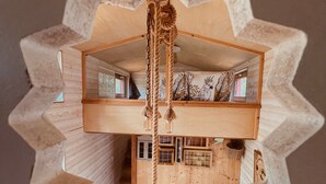 Una cabaña de madera de dos pisos con un dormitorio en el altillo, un columpio de cuerda y un cojín con motivos de pájaros.