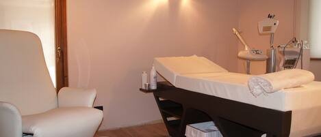 A room with a massage table, a massage chair, a small stool, and a wall-mounted massage machine.