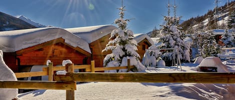 A wooden cabin with a snow-covered roof, surrounded by snow-laden trees and a wooden fence.