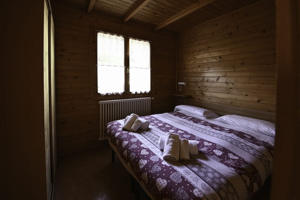 Un dormitorio de cabaña de madera con cama, dos toallas, un radiador y una ventana con cortinas.