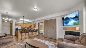 Una sala de estar moderna con un televisor grande, una chimenea y una cocina con armarios de madera y una barra.