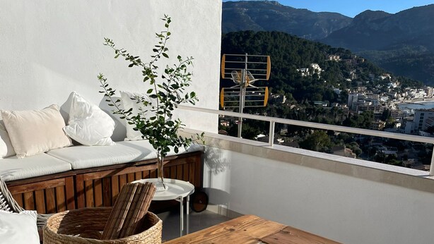 Un balcón con una mesa de madera, cojines blancos y una cesta de mimbre.