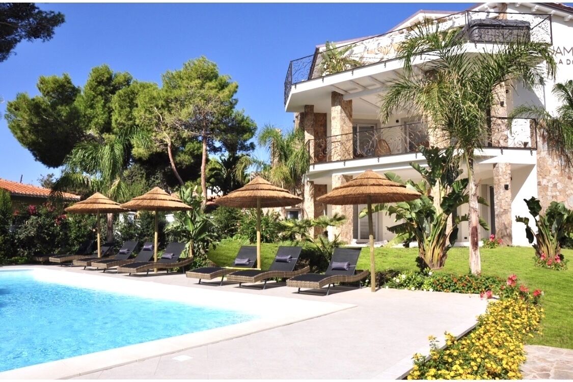 Zona de piscina con sillas de descanso y sombrillas, rodeada de vegetación y un edificio grande con un balcón.