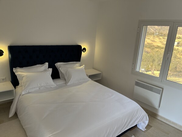 Une chambre à coucher avec un lit, du linge de lit blanc, une tête de lit sombre, des lampes de chevet, une fenêtre donnant sur des arbres et une bouche d’aération fixée au mur.