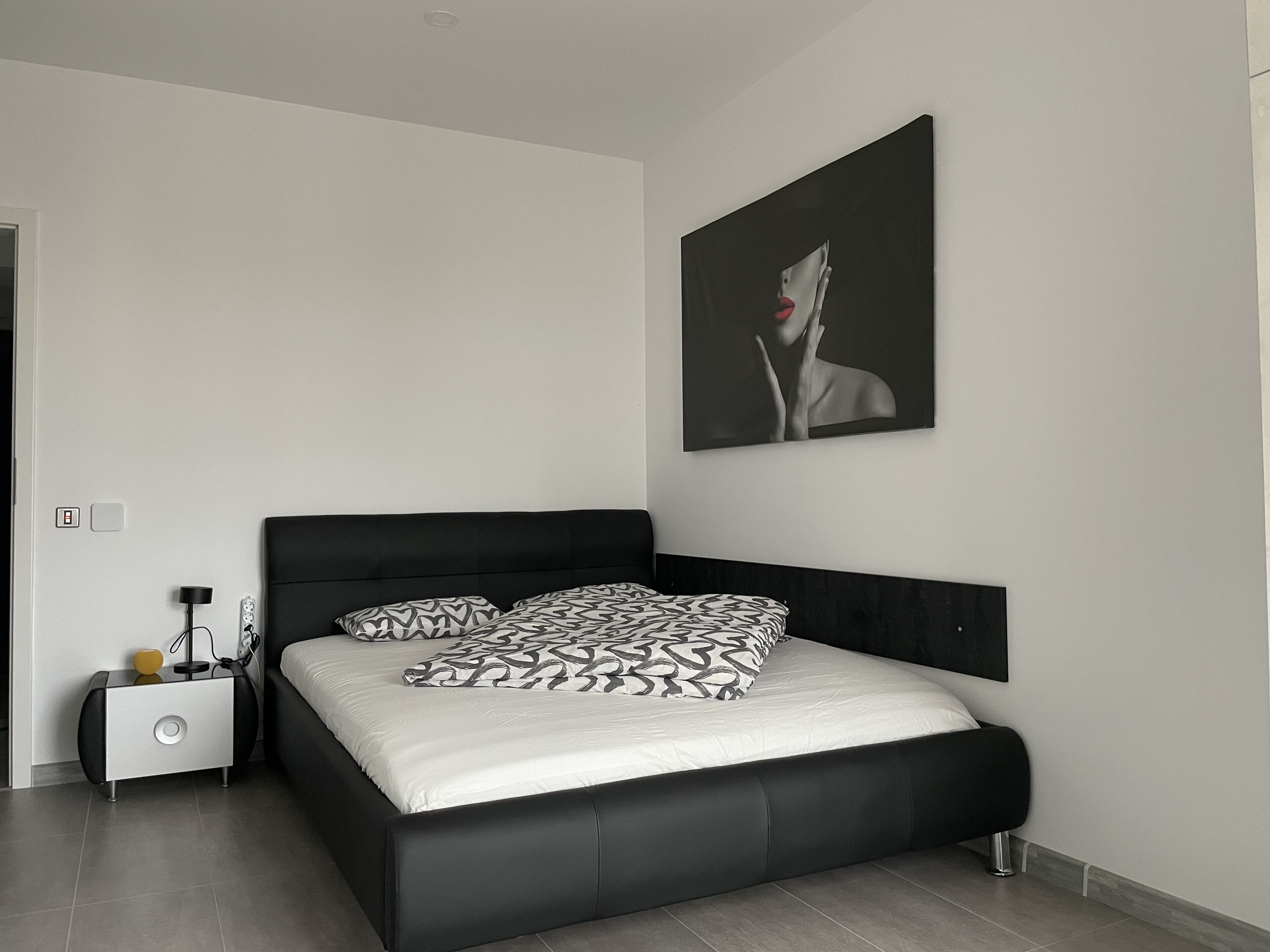 Un dormitorio moderno con cabecera negra, colcha blanca, almohadas estampadas, una mesita de noche con lámpara y una obra de arte colgada en la pared.