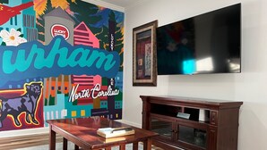 Un salón con una mesa de centro de madera, un mueble bajo de madera oscura, un televisor de pantalla plana montado en la pared y un mural colorido en la pared.