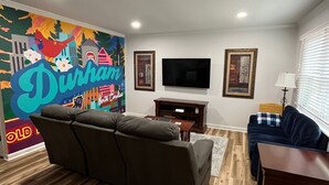 Una sala de estar con un mural grande, un sofá gris, un sofá azul, un televisor y piso de madera.