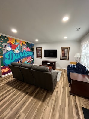 A living room with a large mural, a grey sofa, a blue sofa, a TV, and a wooden floor.