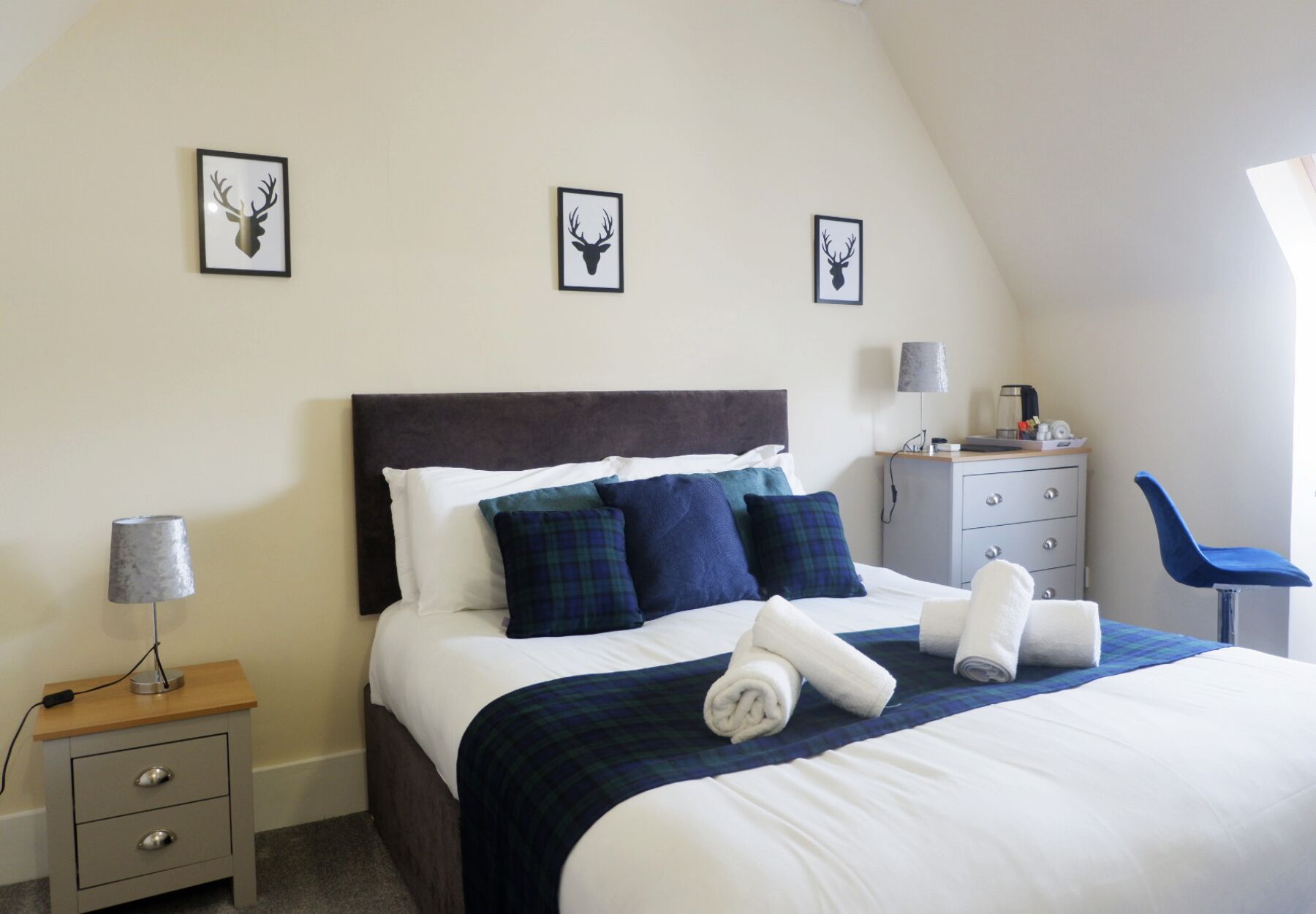 Un dormitorio con una cama, un mesita de noche, un aparador, una silla y tres pares de cornamentas de ciervo enmarcadas en la pared.