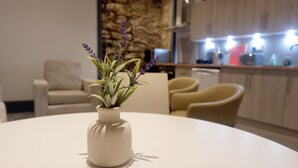 A round table with a white vase containing a plant with purple flowers. In the background, there is a kitchen area with wooden cabinets and a stone wall. - Jacobite house (Inverness)