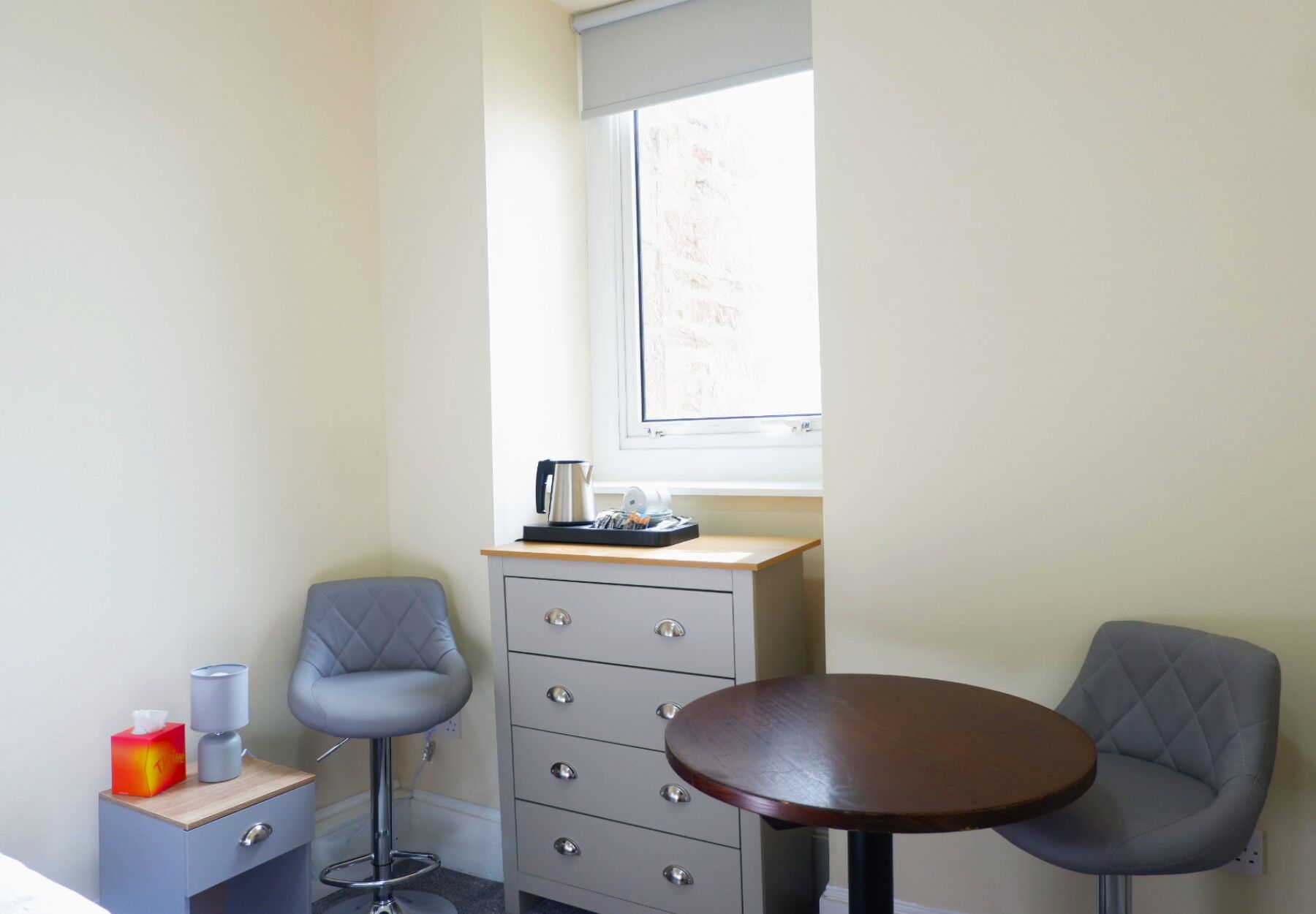 A small room with a single bed, a desk with a chair, a small table with a chair, and a window with a view of a brick wall.