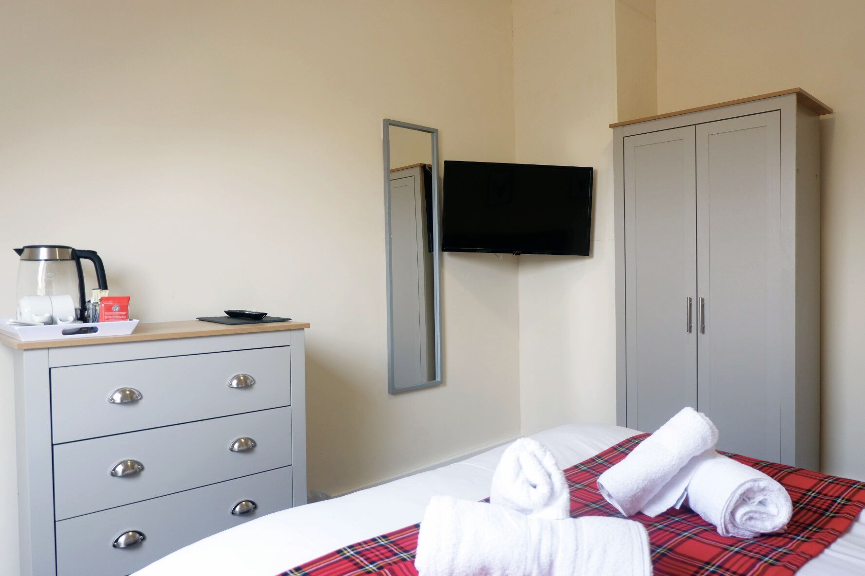 A hotel room with a bed, a grey wardrobe, a television mounted on the wall, and a coffee maker on the bedside table.