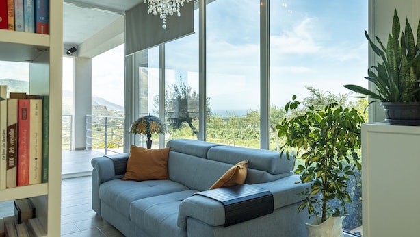 A modern living room with a light blue sofa, a bookshelf, and a view of the outdoors. - Villa Mayas - Scopello with private pool (Castellammare del Golfo)