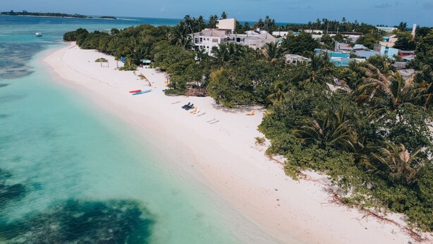 A coastal area with a sandy beach, clear turquoise waters, and a cluster of buildings surrounded by dense tropical vegetation. - Funplace Beach (Himmafushi)