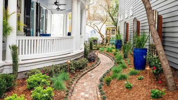 A white two-story house with a curved porch, a brick pathway, and a garden with various plants and a large blue planter. - The Jasmine Suite | Revisit History (Charleston)