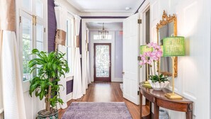 A hallway with a wooden floor, a purple rug, a potted plant, a table with a lamp, and a mirror with an ornate frame. - The Jasmine Suite | Revisit History (Charleston)