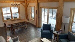 Una habitación amplia con una mesa de comedor de madera, bancos, un sillón azul, un sofá y una pequeña mesa de centro. Grandes ventanales ofrecen vistas de un paisaje nevado.