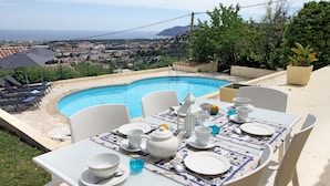 Mesa de comedor con platos, tazones y tetera blancos, con vista a una piscina y al pueblo costero.