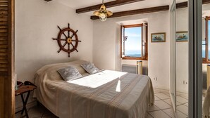 A bedroom with a bed, a ship's wheel decoration, a window with a view, and a door leading to another room. - Pentone - One Bedroom Apartment, Sleeps 4 (Cervione)