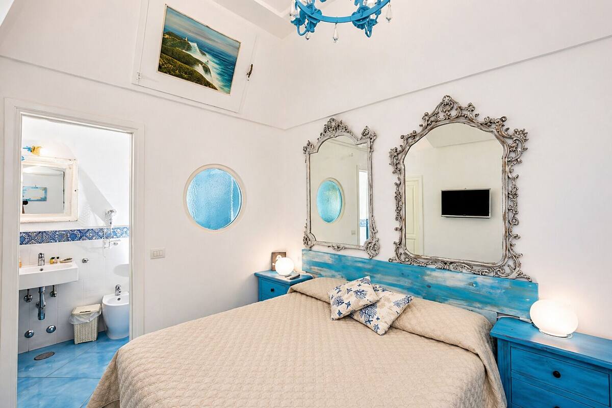 Une chambre à coucher avec un lit, deux miroirs, une table de chevet, une commode, une fenêtre et une salle de bain visible à travers une porte ouverte.